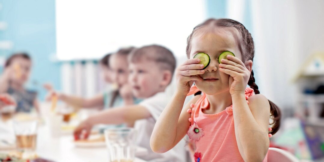 Kindergartenmädchen hält spielerisch Gurke im Gesicht 
