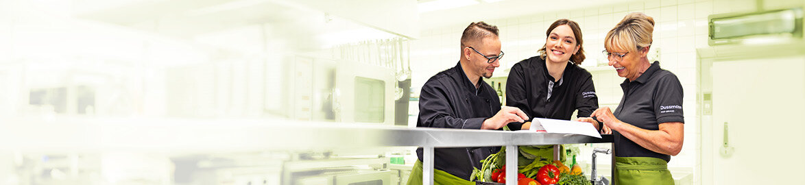 Gruppenbild von drei lächelnden Dussmann-Mitarbeitenden aus dem Gastronomie-Bereich, die in einer Industrieküche stehen und gemeinsam ein Dokument anschauen.