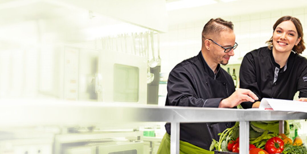 Gruppenbild von drei lächelnden Dussmann-Mitarbeitenden aus dem Gastronomie-Bereich, die in einer Industrieküche stehen und gemeinsam ein Dokument anschauen.