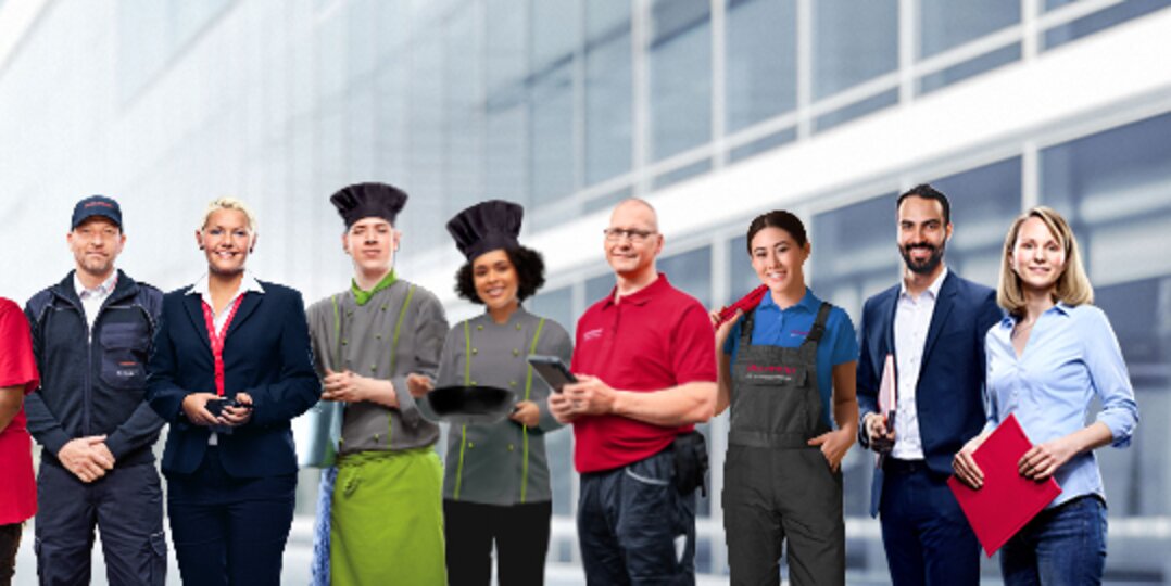 Gruppenbild von lächelnden Dussmann-Mitarbeitenden aus unterschiedlichen Arbeitsbereichen vor neutralem grauem Hintergrund. 